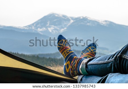 Women's legs in blue jeans and fun socks with a bright yellow pattern lie on the edge of the yellow tent. Mountain ranges covered with green forest and snow. View from the open tent. Royalty-Free Stock Photo #1337985743