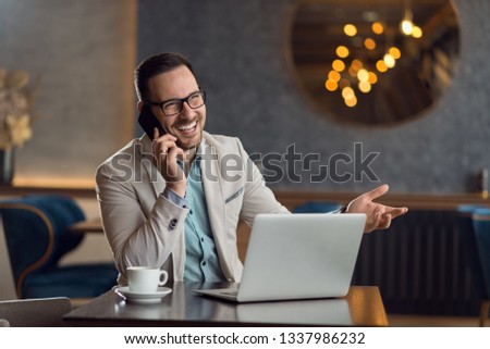 Happy young businessman talking on smart phone while working on laptop in a cafe