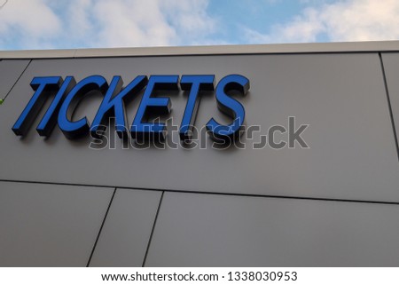 A grey colored exterior wall made of metal composite panels with the word tickets as a sign on the building. The sign is a made of a royal blue glass. There's a blue sky with clouds in the background.
