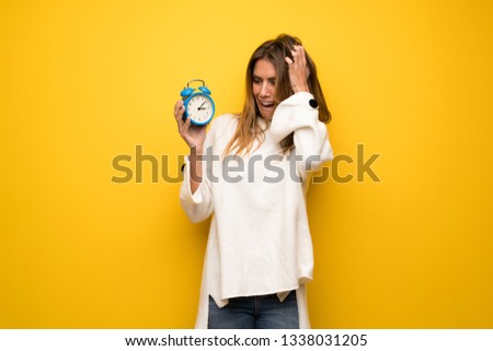 Blonde woman over yellow wall restless because it has become late and holding vintage alarm clock