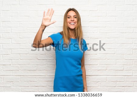 Woman with blue dress over brick wall saluting with hand with happy expression