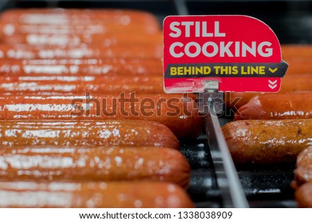 Hot dog wieners cooking on a rolling grill at a truck stop/convenience store with a Still Cooking behind this line sign. 