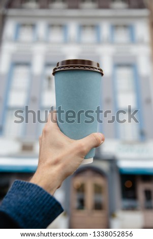 Mockup ad: Male hand holding a blank paper cup in an old town background