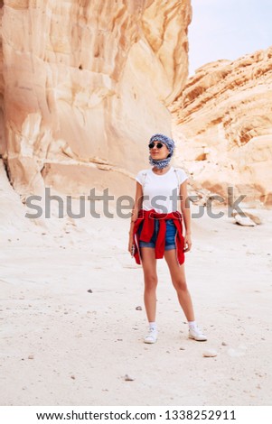 Discovering the secret. Bodacious girl in denim shorts, t-shirt,white sneakers and a kerchief over her head among the sandy rocks and silky blue sky.