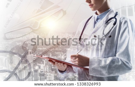 Close up of young woman doctor in white medical suit writing in notebook while standing against city view and medical stuff on background. Double exposure