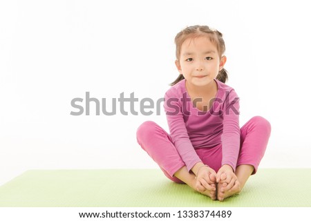 Portrait of cute little Asian girl in purple t-shirt and pants lying face down on green yoga mat, doing yoga exercise and playfully sticking out tongue on isolated white background