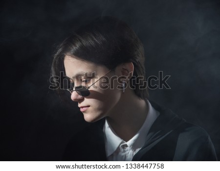 Serious, beautiful girl with short hair sitting in sunglasses under bright light on dark background