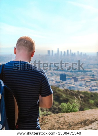 A blond man with a backpack photographed from behind, sitting on a hill and looking at the panorama of Los Angeles in the summer