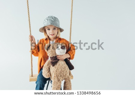 cute kid sitting on swing and holding teddy bear isolated on grey 