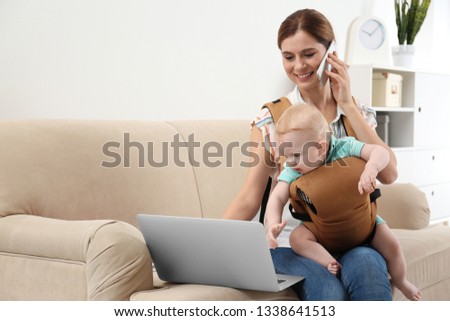 Woman with her son in baby carrier using laptop and talking on phone at home. Space for text