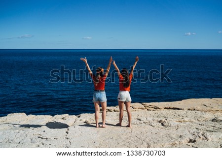 Two young women friends with raised hands, looking at sea. Freedom concept, holiday, beach
