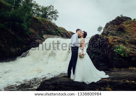 Happy bride and groom stand on the background of the waterfall and hugging.