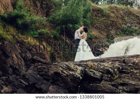 The bride in a luxurious white dress and a beautiful groom hugging against the background of nature. Newlyweds are on a rocky shore, around them the water is boiling.