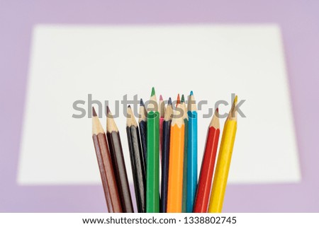 Close-up of a rainbow of colored pencils for child art above the white piece of paper on purple background. 