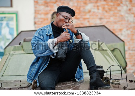 African american man in jeans jacket, beret and eyeglasses, lights a cigar and posed against military armored vehicle.