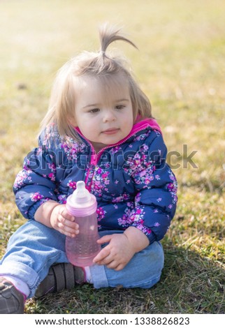 Sweet 17-Months Old Baby Girl Sitting On The Lawn And Drinking Water. Outdoor Photography
