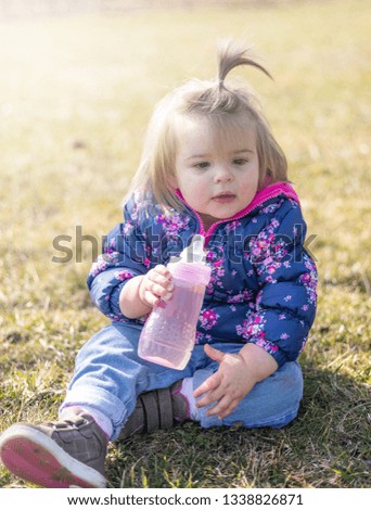 Sweet 17-Months Old Baby Girl Sitting On The Lawn And Drinking Water. Outdoor Photography