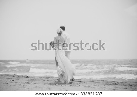 Beautiful young wedding pair of man and woman in white walking along ocean beach shore on windy weather sunny day on blue sky background, horizontal picture