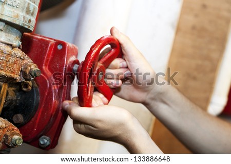 The hands of an adult man in the action of turning a bright red valve, part of an industrial plumbing system.