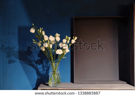 A bouquet of white roses in a glass vase stands against a dark blue wall. Wooden shelves and copy space for text.
