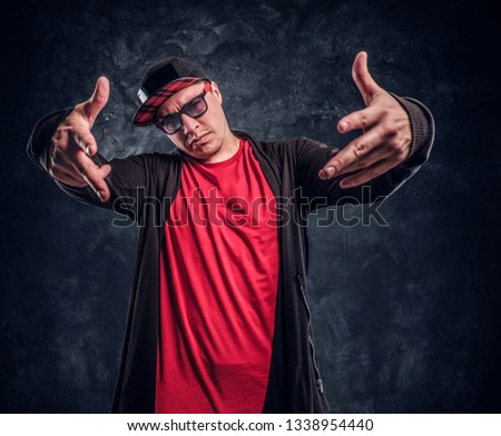 Portrait of a young rapper dressed in a hip-hop style, posing for a camera. Studio photo against a dark textured wall
