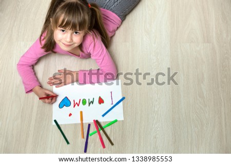 View from above of cute child girl drawing with colorful crayons I love Mom on white paper. Art education, creativity concept.