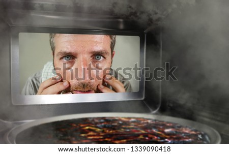 young messy and funny dummy man in the kitchen looking through microwave or oven pizza burning overcooked making a mess of home cook in domestic disaster and lifestyle concept