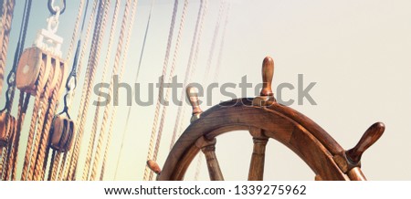 Steering wheel and rigging ropes on an old ship as vintage background for your sea cruise under sails in retro style. Marine equipment with a skipper helm on blur background of ancient sailing vessel. Royalty-Free Stock Photo #1339275962