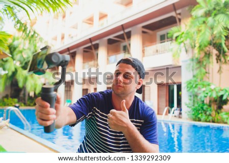Young male video blogger creates video content for his channel. Video shots for users while sitting by the pool. Man vlogger relieves himself on camera with stabilizer.