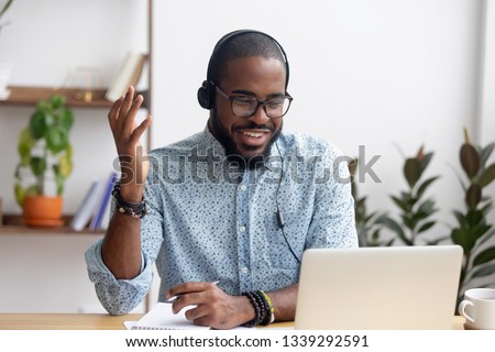 Smiling African American employee in headphones using laptop, looking at screen, making video call or watching webinar, writing notes, distance learning language concept, call center operator working Royalty-Free Stock Photo #1339292591