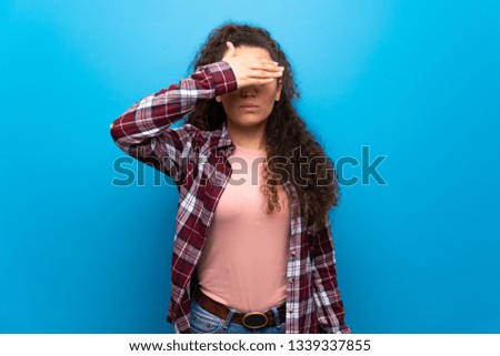 Teenager girl over blue wall covering eyes by hands. Do not want to see something