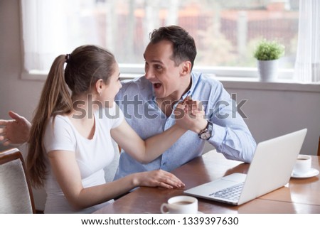 Excited husband holding hand of smiling wife, having good news. Happy millennial couple sitting at laptop. Husband support, happiness, love relationship, success job or family business startup concept