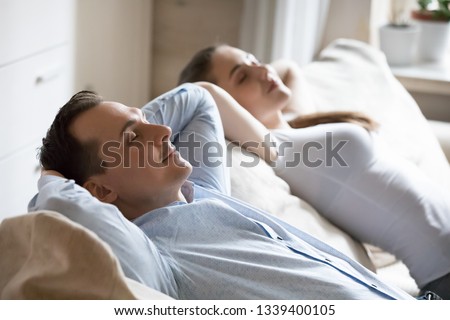 Man and woman relaxing on soft comfortable sofa at home. Happy smiling caucasian couple sitting on couch holding hands behind head enjoying weekend morning together with eyes closed. Stress free image