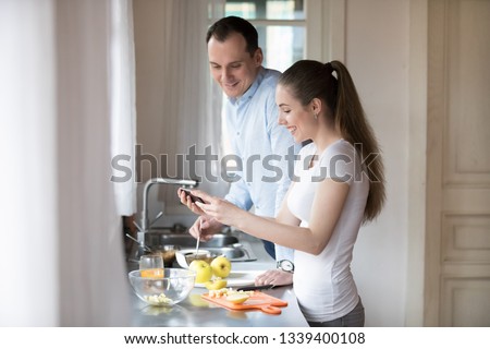 Happy young couple watching video on smartphone while cooking in kitchen at home. Attractive millennial wife laughing showing husband funny clip. Gadget addiction concept. People interacting at home