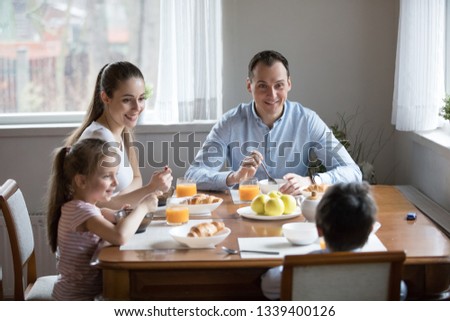 Happy family with children eating morning breakfast at kitchen. Family sitting at dining table at home. Mother and father smiling looking at funny son, daughter laughing brother refuse to eat meal