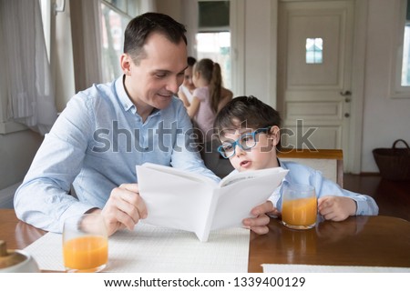 Father and son reading book together at dining room. Family leisure time concept, happy family. Husband and brother making home task while wife and daughter cook breakfast at home on weekend morning
