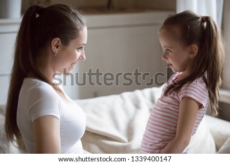 Happy mother looking at smiling daughter holding her hands. Mom and little girl spending time together on weekend at home. Mommy talking to baby. Family leisure time, support, interacting concept
