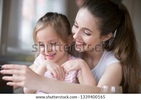 Mother and daughter having fun playing smartphone together. Mommy using gadget entertain little girl, laughing watching funny video on telephone screen. Happy family leisure time, technology concept