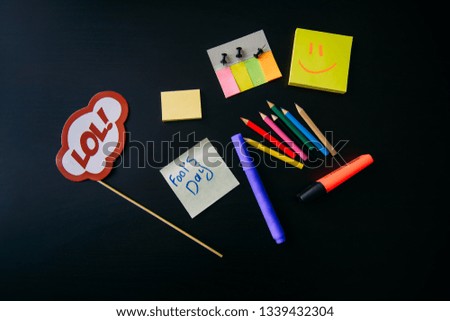April fool's day celebration concept. Office accessories on black background. Sign on a stick. Sticky notes, marker, color pencil.