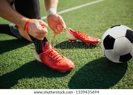 Picture of man lacing soccer sneaker on one leg. He stand on knee. Another sneaker is besides. Ball for game lying on lawn. Close up. Cut view.