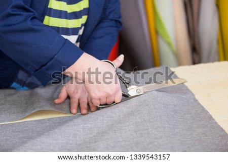 An older woman in a dark coat cuts the marked grey material for the sofa. Behind are different types of materials. She is cutting the material on a wooden desk and preparing it for sewing