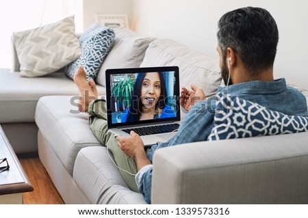 Rear view of husband relaxing on couch while talking to his wife using laptop at home. Mature man lying on sofa and indian woman communicate through video chat on laptop. Royalty-Free Stock Photo #1339573316