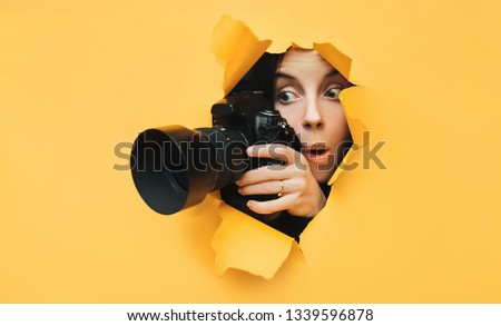 A paparazzi funny girl with a camera looks out from cover and looks at what is happening with her mouth open. Yellow paper, torn hole. Tabloid press. In search of the plot for photo stocks. Royalty-Free Stock Photo #1339596878