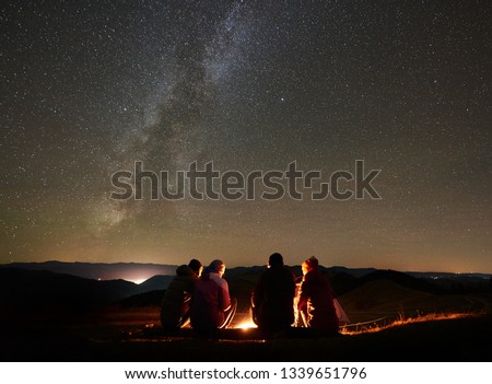 Night summer camping in the mountains. Back view group of four friends tourists sitting on a bench made of logs together around campfire under amazing night starry sky full of stars and Milky way. Royalty-Free Stock Photo #1339651796