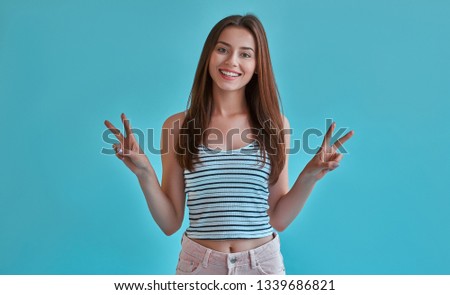 Attractive young woman isolated on blue background. Portrait of happy girl posing, smiling and showing V-signs.