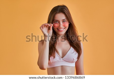 Hello, summer! Attractive young woman in swimwear isolated on orange background. Happy girl in pink sunglasses is posing and smiling.