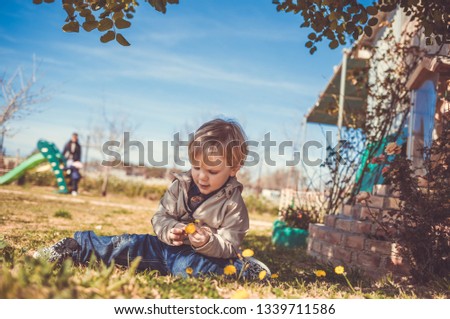 A blond child, two years old, resting on the meadow, sitting in the shade, looks down to the little yellow flowers in his hands. It's a sunny winters morning. A blurred slide is in the background. Set