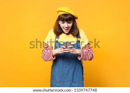 Excited girl teenager in french beret, denim sundress using mobile phone, typing sms message isolated on yellow background in studio. People sincere emotions, lifestyle concept. Mock up copy space