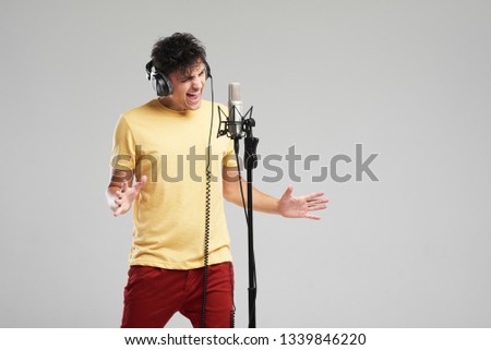 Portrait of rock singer man in studio professional headphones keeping static mic, sings a song loudly on grey background. Concept of rock music and rave