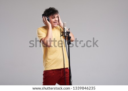 Portrait of rock singer man in studio professional headphones keeping static mic, sings a song loudly on grey background. Concept of rock music and rave